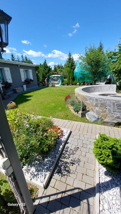 The image depicts a backyard scene on a sunny day. In the foreground, a paved pathway made of gray interlocking bricks leads into the yard. To the left of the pathway, there is a garden bed filled with white gravel and various plants, including flowering bushes with red and pink blooms. To the right of the pathway, a small, neatly trimmed evergreen bush sits on a bed of white gravel.
Further into the yard, a circular, low wall constructed of gray stones surrounds a raised area. The wall has a smooth, light-colored top. A small rock is placed near the base of the stone wall. Beyond this, a well-maintained green lawn stretches across the yard.
To the left of the lawn, a gray, single-story building with white shutters on its windows is visible. In front of the building, potted plants add color. A white trash can and a beige cooler are placed near the building's exterior.
In the background, several evergreen trees and other lush greenery are abundant. A large object covered in green netting is situated towards the right side of the background, potentially a greenhouse or an item under construction. A dark lamppost stands in the far left of the image, casting a shadow. The sky is a clear, vibrant blue with a few scattered white clouds. The overall impression is that of a well-kept residential garden. The image was captured in Zhavoronki, Russia.