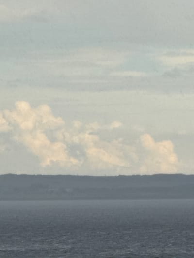 The image shows a body of water in the foreground, a dark, hazy landmass in the middle ground, and a sky with clouds in the background. The sky is a light blue with fluffy white and yellowish clouds. The landmass appears to be a distant mountain range or a very large hill, with layered shadows suggesting depth and undulation. The water is a dark, choppy blue-gray, indicating a potentially rough sea or large lake. There are no people or discernible objects on the water or land. The overall impression is one of a natural landscape, possibly a coastal or lakeside scene, under a partly cloudy sky. The location is identified as Belous, Russia.