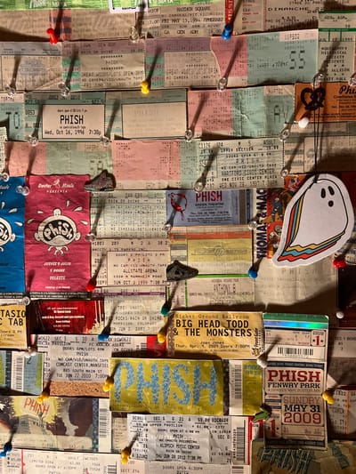 This image displays a dense, vertical close-up view of approximately 50-60 concert tickets, predominantly featuring the band Phish, meticulously pinned onto a light brown, fibrous surface, consistent with a corkboard. The scene is located in the city of Winooski, United States of America.
The tickets vary in color, size, and design, with visible dates spanning from at least 1994 to 2012. Specific dates and venues include October 16, 1996, for "Phish," November 26, 1997, at Hartford Civic Center, October 3, 1999, at Allstate Arena, May 31, 2009, at Fenway Park, and June 27, 2012, at Klipsch Music Center. Other artists and venues mentioned on tickets include "Big Head Todd & The Monsters" on April 4, 2009, "Trey Anastasio" at Red Rocks, and "Anthony B" at Higher Ground.
Numerous pushpins secure the tickets to the surface; these include clear plastic thumb tacks and colored pins in red, blue, yellow, and green, all casting distinct shadows from an implied overhead light source. Interspersed among the tickets are several small decorative items: a white ghost sticker with a rainbow-striped tail on the right side, a dark pretzel-shaped charm near the top right, a colorful miniature shoe charm in the mid-left, a red stylized heart/brain charm with dripping elements in the mid-right, and a dark metallic shoe/boot charm in the lower middle. No individuals are depicted, and the only actions are the static display of pinned objects.