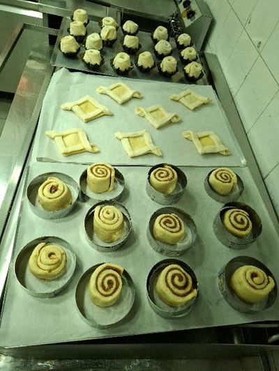The image displays a static scene within a commercial kitchen environment located in Bnei Dror, Israel. Various unbaked pastry items are arranged on baking sheets atop stainless steel preparation surfaces.

In the foreground, a baking sheet covered with parchment paper holds 11-12 individual unbaked spiral pastries, each contained within a circular metal ring mold. These pastries exhibit a light-colored dough with a distinct darker brown, spiraled filling visible from above.

Immediately behind these, on another baking sheet also lined with parchment paper, are 8-10 unbaked square pastries. Each square pastry consists of a flat dough base with its four corners folded inward, creating a smaller, slightly raised central square outline.

In the upper background, positioned on a metal tray, are approximately 18-20 small, unbaked dough pieces, each nestled within a dark brown fluted paper liner. These pieces appear to be gathered or folded, forming a textured, rosette-like shape.

The environment includes reflective stainless steel countertops and a white tiled wall visible on the right side of the frame. An industrial kitchen appliance, possibly an oven or control panel, with dark circular knobs, is partially visible in the upper right background. No individuals are present, and no actions are actively depicted.