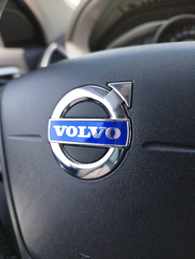 A close-up, eye-level, outdoor, daylight shot focuses on the Volvo logo on the steering wheel of a car. The logo, which is circular with a silver border and a blue insert displaying the word "VOLVO" in silver, is centrally located on the black, textured surface of the steering wheel. The background is blurred, showing parts of the car's interior, including the dashboard and a glimpse of the windshield. The lighting suggests it is daytime, possibly morning or afternoon, with natural light illuminating the scene. The location is not specified in Bremerhaven, Germany, nor are there any people, actions, or interactions visible.