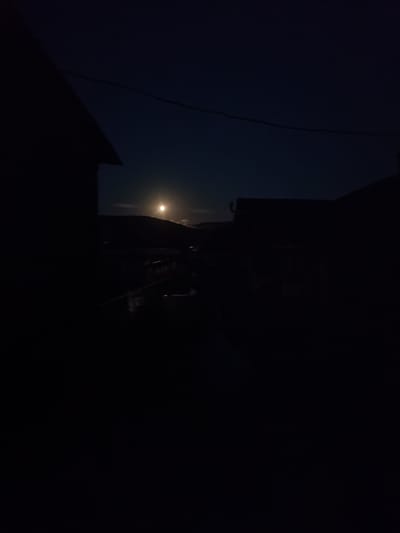 A full moon shines brightly in a dark, clear sky above the silhouette of rolling hills. In the foreground, the dark outlines of several buildings and a power line are visible against the night sky. The scene appears to be a rural or suburban setting in Russia, possibly in the village of Barkhatovo, given the context. The overall mood is serene and quiet, with the moon being the main focal point. No people are visible in the image, and there is no visible text. The time of day is clearly night, illuminated by the bright moon.