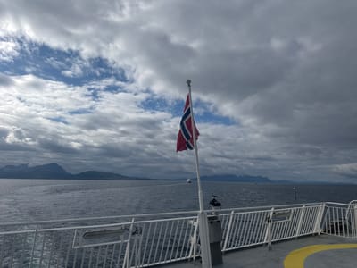 The media file depicts a view from the deck of a ferry at sea, with the Norwegian flag prominently displayed on a flagpole in the foreground. The flag, tattered at the edges, is red with a blue cross outlined in white, symbolizing Norway. The ferry's white railing lines the bottom of the frame, with part of the deck visible on the right, including a yellow painted curve.
The scene extends to a vast expanse of dark, choppy water under a dramatic, cloudy sky. The clouds are a mix of dark gray and lighter patches, hinting at an overcast or approaching storm. In the distance, a chain of rugged mountains rises from the coastline, their peaks partially obscured by the low-hanging clouds, suggesting a mountainous region, likely the fjords of Norway. A few distant boats are visible on the water, including one larger vessel further out.
The overall mood is somewhat somber and vast, characteristic of a journey through a dramatic natural landscape. The time of day is difficult to pinpoint precisely due to the overcast conditions, but the light suggests it is daytime. The location is Lødingen, Norway, which aligns with the coastal mountain scenery and the Norwegian flag. No people are visible in the frame, emphasizing the isolation of the ferry in the expansive seascape.