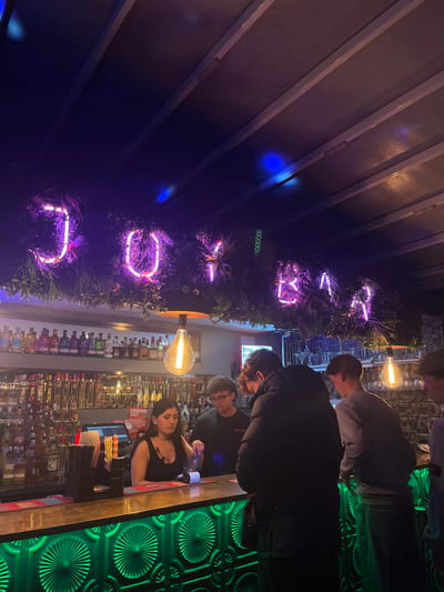 The image shows the interior of a bar in Eastbourne, United Kingdom. The name "Bar Rock" is displayed vertically in neon purple lights against a backdrop of greenery. Several people are standing at the bar, including a female bartender wearing a black shirt who appears to be working. Behind the bar, shelves are stocked with liquor bottles. The bar counter is a light brown color with a red trim. The wall behind the bar is decorated with a patterned green material. Above the patrons, there are vintage-style light bulbs. The ceiling structure is visible in the top-left of the image, displaying a pattern of parallel beams. The image has been rotated 90 degrees counter-clockwise.