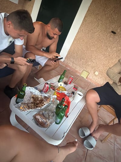 The image depicts a group of individuals gathered around a white plastic table on an outdoor patio. The patio floor is tiled, and a textured, tan wall forms the background. Two men are seated, each holding a mobile phone. One man wears a white t-shirt, while the other is shirtless. A third person, partially visible, is seated on the right, wearing a light-colored top and dark shorts. The table holds several items, including a can of Coca-Cola, a white mug, a bag of bread, and food wrapped in aluminum foil. Green glass bottles are also present on the table. The scene appears to be a casual gathering, possibly a meal or social event, taking place on a warm day. The specific location in Santa Margalida, Spain, cannot be determined from the visual information alone.