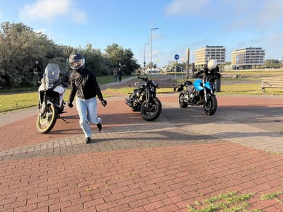 In the city of Hook of Holland, Netherlands, a scene unfolds on a paved walkway with a brick pattern. Two individuals, dressed in motorcycle gear, are positioned near their respective motorcycles.

On the left, a person wearing a black motorcycle jacket, blue jeans, and a black helmet stands next to a white and red motorcycle. The motorcycle has a tall windshield and a yellow rim on the front wheel. This individual is captured mid-stride, appearing to walk away from the motorcycle.

To the right, another person is seated on a bright blue motorcycle. This individual is wearing a white helmet and a black motorcycle jacket. The blue motorcycle is parked parallel to the walkway, facing towards the right side of the frame.

In the background, a black motorcycle is parked in front of a wooden bench. Further back, a paved road is visible with streetlights and signage, including a bicycle symbol. Beyond the road, modern apartment buildings with multiple stories stand under a clear blue sky with scattered clouds. Green trees and bushes line the left side of the scene, creating a natural border between the paved area and the vegetation. The overall lighting suggests it is daytime, with shadows cast by the people and motorcycles on the walkway.