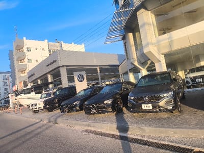 The image captures an urban street scene in Gonyeli, Northern Cyprus, under the clear sky of what appears to be late afternoon or early evening, characterized by warm light and long shadows cast by the vehicles.

The main subjects are a series of high-end vehicles parked along a paved curb on the side of an asphalt road. These vehicles are positioned in front of a modern commercial building, which houses a car dealership. The dealership building features large glass display windows and a prominent circular Volkswagen logo on its facade. To the right of the dealership, another contemporary building with angular architectural elements and reflective panels is visible. Behind the dealership, a multi-story residential building with balconies can be seen.

From right to left, the parked vehicles include a grey Volkswagen T-Roc SUV, which has "T-Roc" clearly printed on its front license plate. Next to it is a black Mercedes-Benz sedan, featuring an "SKC" sticker on its front bumper. Further down the line, a black Mercedes-Benz G-Class SUV is followed by a white Mercedes-Benz G-Class SUV. Partially visible behind the white G-Class is a large white boat or yacht, suggesting a diverse range of merchandise or nearby marine facilities.

The scene depicts a commercial area where vehicles are likely on display or parked for sale. No people are clearly visible engaging in any specific activity. Utility lines are faintly visible against the bright blue sky. The overall atmosphere is bright and commercial.