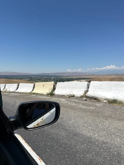 The media file, a photograph taken from inside a vehicle, depicts a scenic view of a valley in Беш-Мойнок, Kyrgyzstan. The perspective is from the passenger side, with the side mirror prominently featured. The mirror reflects a portion of the sky and what appears to be a car's interior, possibly a person's arm and shoulder.
The scene outside the vehicle showcases a vast, clear blue sky dominating the left side of the frame. To the right, a roadside barrier, constructed from large, whitewashed blocks, runs along a paved road. Beyond the barrier, the landscape unfolds into a valley. This valley features rolling hills with dry, earthy tones, dotted with patches of green vegetation and signs of human habitation, possibly a small town or scattered buildings. The terrain suggests a mountainous or hilly region, characteristic of Kyrgyzstan.
The time of day appears to be late morning or early afternoon, given the bright sunlight and clear sky. The weather seems fair and dry. The white barrier is weathered, with some cracks and signs of dirt accumulation, indicating it has been in place for some time. Rust is visible on the white trim of the car's body in the foreground, adding to the sense of ruggedness or age of the vehicle.
The composition suggests a journey or a stop along a scenic route. The view through the side mirror offers a secondary perspective, hinting at the presence of the photographer or another passenger. The overall impression is one of a quiet, perhaps remote, natural landscape, viewed during a road trip. There is no visible text in the image.