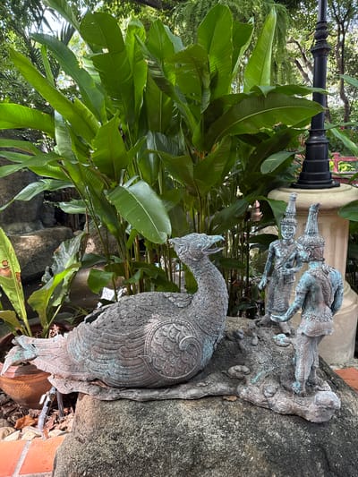 The image depicts a garden setting in Samrong Nuea Subdistrict Municipality, Thailand. A bronze-colored statue of a bird, resembling a peacock, is positioned in the foreground. The bird is highly detailed, with intricate patterns on its feathers. Behind the peacock statue, there are two bronze-colored statues of male figures wearing traditional Thai headdresses. These figures appear to be positioned on a rock or stone structure. Lush green foliage, including large, broad leaves, fills the background, obscuring some of the statues and structures. A black lamppost is visible among the leaves. The statues are situated on a textured stone surface with a mix of natural elements like rocks and plants. The overall impression is that of a serene, landscaped garden with culturally significant sculptures.