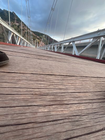 The image depicts a close-up perspective of a wooden pedestrian bridge extending across a valley in Tabaroucht, Morocco. The planks of the bridge are visible in the foreground, running parallel to the frame, showing a slightly worn, weathered surface. A portion of a shoe is visible in the upper left corner of the image. To the right of the wooden walkway, a metal support structure with a repeating triangular pattern runs the length of the bridge. Suspension cables extend upwards from the bridge to the sky. In the background, a hillside covered with sparse vegetation is partially visible, meeting the sky. The sky is overcast with gray clouds.