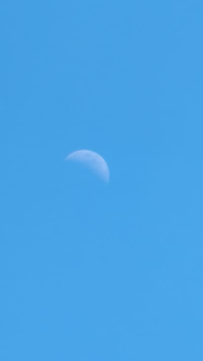The image shows a partial view of the moon against a clear blue sky. The moon is illuminated on the right side, with a crescent shape visible. The sky is uniformly blue with a slight texture of small, speckled dots that could be noise from the image capture or atmospheric particles. There are no discernable landmarks or elements that would suggest a specific location in Kozare, Bulgaria, or any other city. The scene is entirely natural, focusing on the celestial body and the sky.