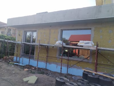 This image captures a construction site in Dumbraveni, Romania, likely in the late afternoon given the warm, diffused lighting. The scene depicts the exterior of a building under renovation or construction, with scaffolding erected around the lower section. The wall is covered in yellow insulation panels, partially secured with grey adhesive. A blue layer is visible at the base of the wall, possibly a moisture barrier or primer. Large windows with reflective glass are installed, offering a view of the surrounding environment, including a red-tiled roof of another building and trees.

In the foreground, the ground is a mix of dirt and construction debris. Several rolls of black material, possibly roofing felt or similar construction supplies, are stacked on the right side of the image. Some pieces of yellow foam insulation are scattered on the ground. Two white bags of insulation are placed on the scaffolding, one clearly displaying the brand "NOVOTERM" with the text "basalt insulation."

The overall impression is one of ongoing construction work. The sky is bright and clear, suggesting fair weather. There are no people visible in the image, focusing solely on the building and its immediate surroundings. The setting appears to be a semi-urban or rural area, typical of residential construction in Romania.