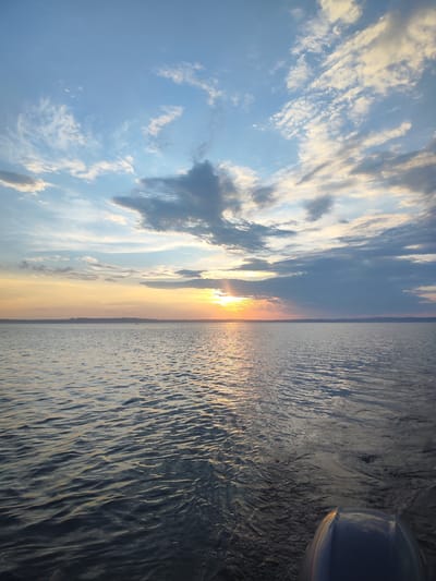 A wide-angle, eye-level shot captures a serene sunset over a large body of water, likely a lake or calm sea, in Galevo, Russia. The sky is a dynamic canvas of blues, oranges, and grays, with scattered clouds catching the warm glow of the setting sun. The sun itself is a bright, diffused orb peeking through a bank of clouds near the horizon, casting a shimmering golden path across the water's surface. The water is calm, with gentle ripples reflecting the sky's colors, and appears dark in the foreground, transitioning to a brighter, more reflective surface where the sun's rays hit. The distant shoreline is a faint, dark silhouette under the vibrant sky, suggesting a landmass or treeline. In the far distance on the water, a tiny dark speck may indicate a small boat or vessel. The lower right corner of the image shows the curved, dark surface of what appears to be part of a boat's hull or a similar structure, suggesting the photo was taken from a watercraft. The overall atmosphere is peaceful and expansive, emphasizing the vastness of nature.