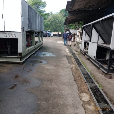 The image shows two large industrial cooling units, likely air conditioners, parked outside in a paved area. The units are located in the city of Kecamatan Curug, Indonesia. One unit is partially obscured by a covered structure on the right. The ground is wet, with puddles of water. A man and a woman are walking away from the camera, toward the right side of the image. The man is wearing a dark blue shirt and jeans. The woman is wearing a pink shirt and blue jeans. Other individuals are visible further down the paved area, but they are too far to make out details. Both cooling units are made of metal with louvered sections. Several vehicles are parked in the background. The scene is a typical industrial area, likely a factory or manufacturing plant.