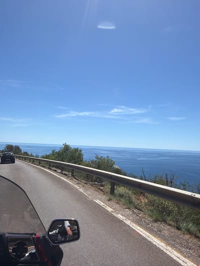 The image shows a view from a motorcycle traveling along a coastal road. The road is paved and curves to the left, with a metal guardrail along the right side. Beyond the guardrail, there are green trees and shrubs. The road overlooks a vast expanse of blue ocean, with a few small whitecaps visible. The sky is a clear, bright blue with some wispy white clouds. In the foreground, a portion of the motorcycle's windshield and a side mirror are visible. The side mirror reflects the image of a person's arm wearing a black glove and a white and yellow shirt. A dark colored car is seen on the road ahead. The scene appears to be in a sunny, coastal location.