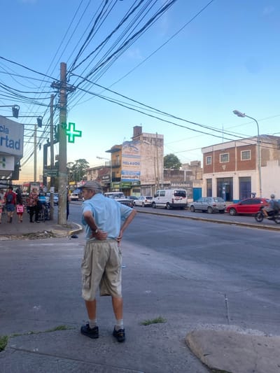 The image presents an urban street scene in Libertad, Argentina, under a clear light blue sky. In the foreground, an elderly male is positioned on a concrete sidewalk, facing away from the viewer and slightly to the right. He wears a light blue short-sleeved collared shirt, khaki cargo shorts, light-colored socks, and dark athletic shoes, with both hands placed on his lower back. To his left, on the same sidewalk, other pedestrians are visible, including an adult and a child in pink clothing.
A prominent utility pole, densely covered with multiple black overhead utility wires, stands behind the foreground male and to his left. This pole features a bright green, illuminated cross, indicative of a pharmacy. A partially visible sign to the far left reads "Farmacia Libertad."
Across the asphalt street, multiple vehicles are parked along the right curb, including a white vintage pickup truck, a white cargo van, a silver hatchback, and a red sedan. A motorcyclist on a dark motorcycle is in motion on the street, traveling leftward, visible behind the red sedan. Buildings line the street opposite the foreground sidewalk. Notable structures include a multi-story building displaying a yellow and brown banner advertising "La Tradicion HELADON," and another building to its right with a brick upper facade and a ground-level sign reading "ABOGADOS."