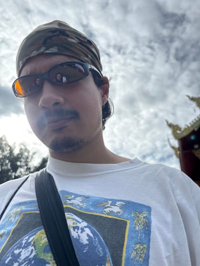 A male subject is depicted in a low-angle, close-up outdoor photograph in Ban Mae Na Chalong, Thailand. The subject has dark hair, a mustache, and a short goatee, and wears a camouflage-patterned bandana. He wears dark, wraparound sunglasses with amber-tinted lenses, reflecting two individuals; one clearly holds a white smartphone, indicating a photographic interaction. The subject wears a white t-shirt with a graphic print depicting the Earth, pelicans, and palm trees, and a dark strap (likely from a bag) crosses his chest. An earring is visible in his left ear. The background on the subject's right features ornate golden Thai temple architecture, including a multi-tiered spire. Out-of-focus green foliage is visible to the subject's left. The sky is overcast with scattered white and grey clouds.