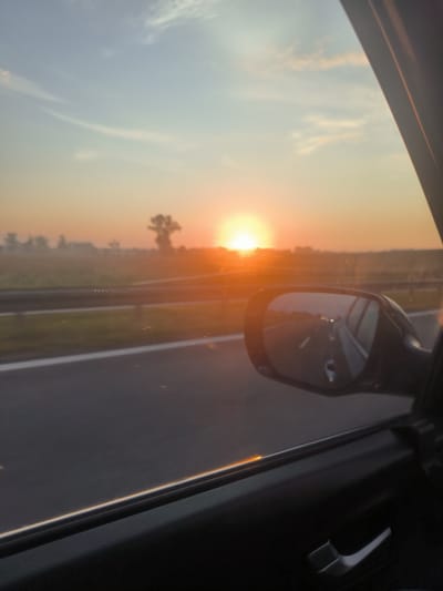 The image depicts a sunset viewed from inside a car traveling on a highway near Działosza, Poland. The sun, a bright orange orb, is low on the horizon, casting a warm glow across the sky and landscape. In the foreground, the interior of the car is visible, including a car door, window frame, and a side-view mirror reflecting the road ahead and the surrounding scenery. The highway stretches into the distance, bordered by fields. A solitary tree stands silhouetted against the vibrant sunset sky. The overall impression is one of a peaceful journey during golden hour.