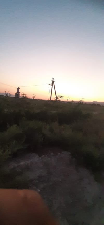 The image depicts a rural landscape at dusk. In the background, a hazy sunset paints the sky in gradients of blue, pink, and yellow. Silhouetted against the horizon are rolling hills. In the mid-ground, two utility poles stand amidst sparse vegetation and a larger, more distant pole with insulators. The foreground is dominated by dark, dense shrubbery and a patch of rocky ground, with a blurred, orangish object partially visible in the bottom left corner, likely a hand or finger. There are no visible people or specific landmarks to indicate the city of Сафедорон, Tajikistan.