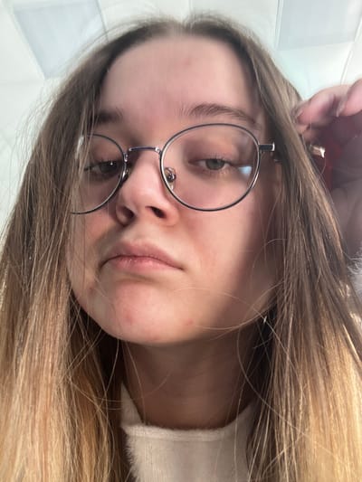 The image displays a close-up, head-and-shoulders portrait of a young individual, likely female, with fair skin and long, light brown hair with blonde highlights, parted centrally. The individual wears silver-rimmed, round spectacles, through which light-colored eyes are visible, looking slightly downwards. The facial expression is neutral. Minor reddish blemishes are present on the forehead and chin. The individual's left hand (viewer's right) is partially visible near the right side of the head, appearing to touch or adjust hair or the glasses' earpiece. A section of a light-colored, possibly white or cream, soft-textured garment is visible around the neck and chest area. The background is indistinct and appears to be an indoor setting, with a portion of a white ceiling featuring rectangular panels or tiles visible at the top. The image does not contain any visual information that allows for the identification of a specific location within the city of Большевик, Russia.