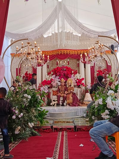 A wedding ceremony is taking place under a white tent, decorated with red drapes and ornate chandeliers. In the center of the raised stage, a newlywed couple, dressed in traditional attire, is seated on a white sofa. The bride wears a golden headpiece and a matching intricate gown, while the groom is adorned in a maroon suit with gold accents. They are surrounded by a profusion of red and pink flowers. Two large, golden, heart-shaped arches frame the stage, adorned with hanging crystals. In the foreground, a red carpet leads to the stage, with floral arrangements flanking the sides. Several people are visible in the background, some attending the ceremony and others involved in its setup. The overall ambiance suggests a festive and opulent celebration.