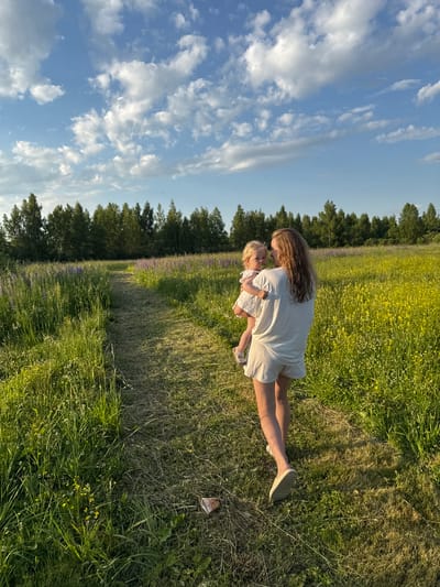 A woman carries a young child through a grassy field in the village of Koloksha, Russia, on a clear, cloudy day. The woman, facing away from the camera, wears a light-colored outfit consisting of a t-shirt and shorts. She cradles the child, who is also in light-colored clothing and has a pacifier in their mouth. A mown path runs through the field, bordered by tall green grass and scattered yellow and purple wildflowers. In the distance, a dense line of trees forms a horizon, with the bright blue sky and scattered white clouds above. The image captures a serene moment of a mother and child enjoying a walk in nature.