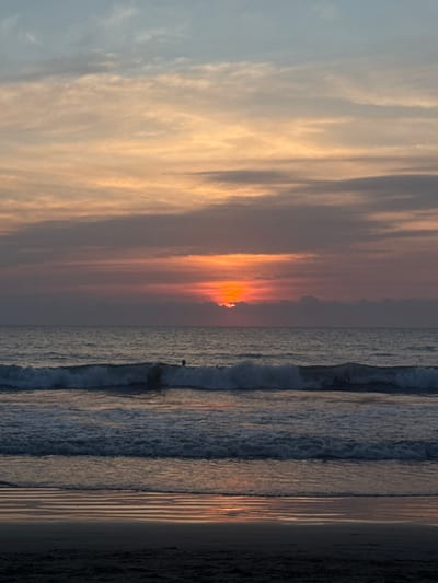 The image shows a sunset at the beach. The sky is filled with clouds, and the sun is setting on the horizon. The colors of the sunset are reflected in the ocean. The waves are crashing on the shore, and the beach is sandy. A person is standing in the ocean looking at the sunset.