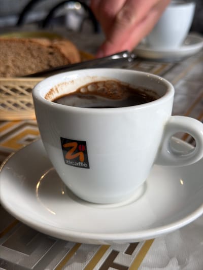 A close-up, eye-level shot focuses on a white ceramic coffee cup filled with dark coffee and topped with a layer of crema. The cup is positioned on a matching saucer, both resting on a table with a patterned tablecloth. To the left of the cup, a basket containing slices of brown bread is visible. In the background, out of focus, a person's hand is seen reaching towards another out-of-focus white coffee cup and saucer. The image appears to be taken indoors, likely in a cafe or restaurant setting. There is no information in the image that suggests the location is the city of Kosteneevo, Russia.