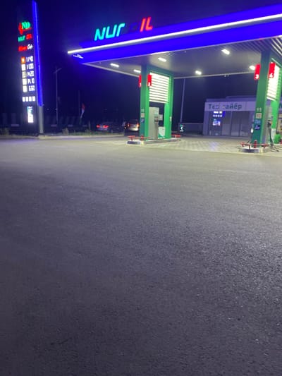 The image captures a nighttime view of a Nur Oil gas station in Shahrinav, Tajikistan. The station features a prominent blue canopy illuminated by white lights, with the Nur Oil logo displayed in neon green letters. To the left, a tall illuminated sign displays prices for various fuel types, including "11.40", "11.00", "10.20", "10.10", and "6.50". Several cars are visible in the background, parked or in the process of refueling. A small building with the text "Тел: майер" is situated behind the pumps. The ground is paved with asphalt, and the overall scene is lit by the gas station's artificial lighting, creating a vibrant and colorful nighttime atmosphere.