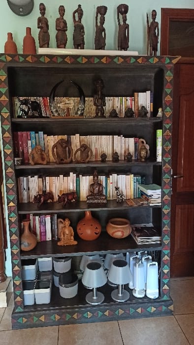 A cabinet featuring cultural artifacts and modern objects. Left shelves display carved wooden figures and books with wooden sculptures. The middle shelves contain stacked books adorned with various carvings, including animal figures. The right shelves showcase pottery-like items alongside a set of modern, cylindrical objects stacked with chrome bases. A triangular patterned border frames the cabinet's front.