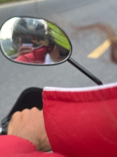 A close-up shot focuses on the left side-view mirror of a vehicle, likely a motorcycle or scooter, in Vị Xuyên Commune, Vietnam. The mirror has an oval shape with a metallic rim, and its dark stem extends diagonally towards the upper right.
To the right of the mirror, a person's arm is visible, dressed in a vibrant red garment that features a distinct vertical white stripe along its side. Below the sleeve, a light-skinned hand firmly grips a dark, possibly black, handle or control, suggesting the person is operating or about to operate the vehicle.
The surface of the mirror reflects a blurred scene: indistinct green shapes, likely foliage or greenery, are prominent, along with hints of red and darker, shadowy forms, possibly another person or the person's own reflection. The background behind the mirror and the arm is uniformly blurred in shades of gray, consistent with a road surface or asphalt. In the upper left quadrant of the image, a bright, diffused yellow shape suggests a light source or a reflected object. The scene appears to be outdoors during daytime. No text is visible in the image.