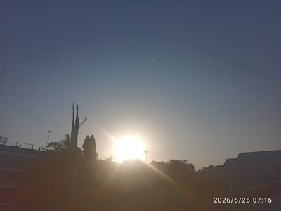 The image captures a sunrise over a cityscape, with the sun prominently displayed in the center, casting bright rays across the frame. The sky is a clear, pale blue, transitioning to a lighter hue near the horizon. Silhouetted against the bright sun are trees, buildings, and what appears to be a barbed-wire fence along the top of a wall. The overall scene suggests an urban environment in Embakasi, Kenya. The date and time stamp "2026/6/26 07:16" is visible in the bottom right corner of the image. There are no people visible in the frame.