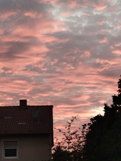 The image depicts a dramatic sunset sky filled with wispy clouds tinged in shades of pink and gray, extending over the rooftops of Kitzingen, Germany. In the foreground, the upper portion of a house is visible, featuring a dark red tiled roof with a chimney and a satellite dish. Adjacent to the house, silhouetted trees partially obscure the view, with a few branches extending into the frame. The lower part of the house shows a window with closed blinds. The scene is devoid of any visible people or animals.