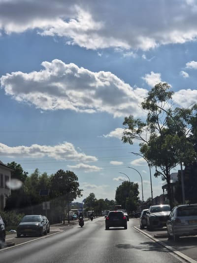 This is a vertical, eye-level, medium shot of a road in Käerjeng, Luxembourg, under a partly cloudy sky. The foreground features a wet asphalt road with tire tracks, suggesting recent rain. Several vehicles are present: a dark SUV on the right side of the road with its license plate visible, a silver sedan parked on the right, a dark hatchback in the center lane, and a motorcycle in the left lane. The background shows trees lining the road, a building on the left, and streetlights. A gas station with a sign reading "RVS" is visible in the distance. The sky is blue with large, white cumulus clouds and thinner, elongated clouds. The overall impression is a typical day in a suburban or semi-urban environment.