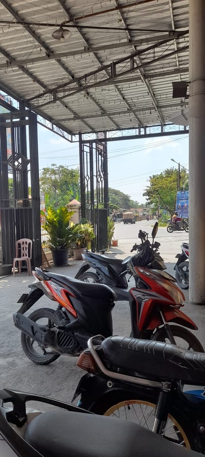 In Crewek, Indonesia, a paved outdoor area is covered by a corrugated metal roof supported by metal beams and columns. Beneath the roof, several motorcycles are parked. In the foreground, the rear of a black motorcycle is visible, with a tan seat and a yellow rim on the rear wheel. To its left, an orange and black scooter is parked, facing left. Behind it, a black scooter is also parked, facing left. A light pink plastic chair sits to the left of the scooters, near a tall potted plant with large green leaves. Beyond the motorcycles, a street is visible with traffic, including a truck carrying goods and a person riding a motorcycle. Trees line the street, and a blue sign with text and images is mounted on a pole. The overall scene suggests a roadside or open-air parking area in a populated area.