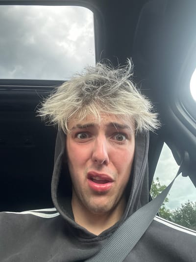 The image shows a close-up view of a young man's face. His hair is white and messy, and he is wearing a black hooded sweatshirt with white stripes and a black seatbelt fastened across his chest. He appears to be sitting in a car, looking at the camera with a shocked expression. His mouth is slightly open, and his eyes are wide. The sky is visible through the sunroof of the car, and there are a few leaves visible through the car window. The location is Arkendale, United Kingdom, coordinates (latitude: 54.052, longitude: -1.387).