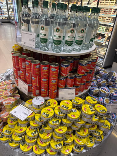 A multi-tiered circular display in a store holds various canned goods and bottled spirits. Clear glass bottles with green accents, labeled "MINI ΟΥΖΟ ΜΥΤΙΛΗΝΗΣ", occupy one section. Red cans marked "CALIFORNIA SQUID" and "FLOKOS ΚΑΛΑΜΑΡΙ" fill another, alongside smaller cans of "ZANIA". Yellow-labeled cans with black writing, likely containing stuffed grape leaves ("ΝΤΟΛΜΑΔΑΚΙΑ ΓΙΑΛΑΝΤΖΙ") dominate another part of the display. Price tags are visible, with one reading "11,20€" and another "4,67€". The arrangement suggests a curated selection of Mediterranean foods and spirits.