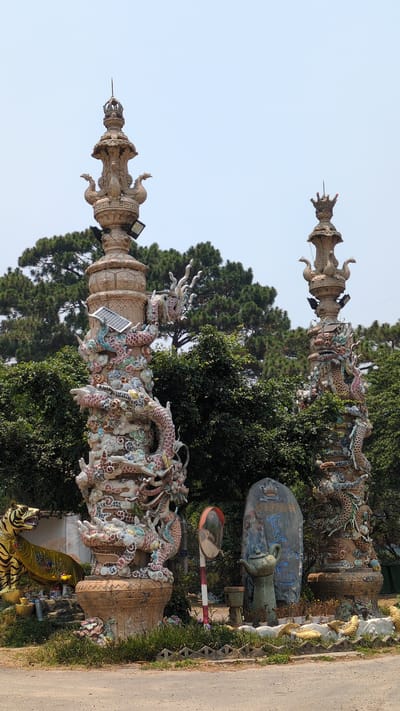 The image shows two ornate pillars in Lâm Sơn, Vietnam. The pillars are decorated with colorful dragon sculptures and topped with spire-like structures. A solar panel is attached to the side of one pillar. Lush greenery and trees surround the pillars, and a road is visible on the right side of the frame. Other statues and objects are visible in the background, including a large stone monument and a traffic mirror. The sky is pale blue.