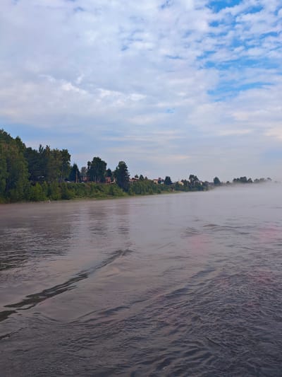 An overcast sky with scattered white clouds over a wide, calm river. A bank of trees and houses lines the left side of the river, with a dense forest in the background. A light fog or mist hovers over the water in the distance. The foreground shows ripples and waves on the river's surface. The scene depicts a quiet, rural landscape.
