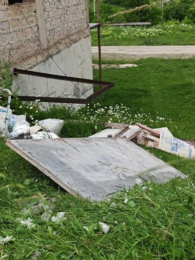 This media file shows a close-up, low-angle view of construction materials and debris scattered in a grassy area next to a partially constructed building. In the foreground, a large, flat piece of gray material, possibly metal or concrete, lies at an angle on the grass. Behind it, several white sacks, some with visible text indicating they contain cement and are for export, are piled up. Also visible are pieces of wood and other debris.

The building's foundation is made of concrete, and its upper wall is constructed with rough-hewn, light-colored stones. A dark metal beam extends horizontally from the building. The scene is set outdoors with lush green grass and scattered white wildflowers. In the background, a dirt road or pathway is visible, bordered by more greenery and trees. The overall impression is one of an unfinished construction project in a rural or semi-rural setting, possibly in Gugark, Armenia. The lighting suggests it is daytime, and the weather appears to be fair, conducive to outdoor work or for materials to be left outside. No people are visible in the frame, and the overall mood is one of a work in progress, with a touch of natural beauty from the wildflowers.

Visible text on the sacks includes: "ORY AND C", "PEM", "EXPORT", "PORT", "TRAN", "FOR EXPORT", "PEM-500", "LANDMENT", and "MIA CEMEN".