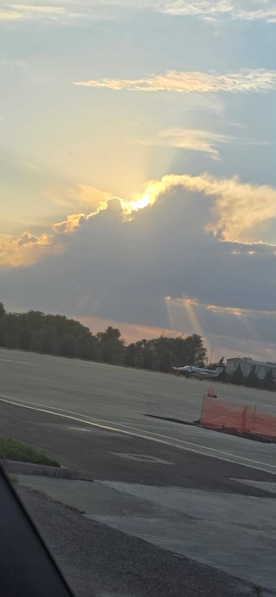 The image depicts an airfield scene at sunset in Dachi Su, Kyrgyzstan. The sun is partially obscured by large, dramatic clouds, casting crepuscular rays through the sky. In the foreground, a dark-colored vehicle occupies the lower left portion of the frame. The middle ground features a paved airfield surface with distinct markings, a line of trees forming a dark border, and a small airplane positioned on the tarmac. Further back, several buildings are visible, and a section of orange construction fencing is present. The sky is a blend of pale blue and the warm hues of sunset, with scattered clouds.