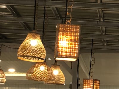 The image shows a cluster of four woven lamps hanging from a ceiling structure. The lamps are made of a light-colored natural material, likely bamboo or rattan, and emit a warm, inviting glow from internal lights. Two of the lamps are bell-shaped, with intricate woven patterns that create a textured effect. One is a rectangular lantern-style lamp, also woven, with a grid-like pattern. The fourth lamp, partially visible on the far left, appears to be similar in shape to the bell-shaped ones. The ceiling above is industrial in appearance, with exposed metal beams and corrugated panels. The overall impression is one of a cozy, possibly rustic or bohemian interior space, perhaps a restaurant, cafe, or home, with a focus on warm lighting and natural materials. The time of day is likely evening or night, given the prominent lighting from the lamps and the dim ambient light. The location context of Blangbitra, Indonesia, suggests a tropical or subtropical setting, which aligns with the use of natural, woven materials in the decor. There is no visible text in the image.