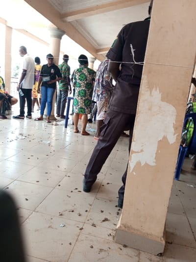 The scene depicts a group of individuals gathered within a covered outdoor area, possibly a veranda or open hall, in Ajalli, Nigeria. The area features a light-colored tiled floor and multiple beige support columns with visible flaking paint or plaster.
In the immediate foreground on the right, the lower body and back of a person wearing a dark brown/black short-sleeved uniform shirt and cargo pants are partially obscured by a column. A white string or wire runs horizontally across the upper back of this individual and extends across the scene.
In the midground, approximately seven to eight individuals are standing. From left to right: a person in a white short-sleeved shirt and dark pants holds a clear plastic bottle; a woman in a black t-shirt, blue jeans, and a black baseball cap holds a white rectangular object; another person wears a green, yellow, and black patterned dress; other individuals wear various patterned clothing, some partially obscured by columns or other people. A blue plastic stool is visible between some individuals.
The tiled floor is notably scattered with numerous small, dark, irregular objects, possibly debris, dirt, or discarded items. Bright ambient light indicates daytime.