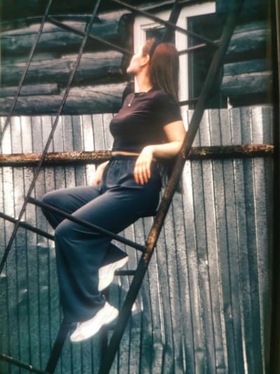 The media file depicts a young woman with long, dark hair, wearing a dark t-shirt and dark, loose-fitting pants, and white sneakers. She is sitting on a metal ladder that is positioned against a corrugated metal wall, possibly a fence or part of a building. The ladder appears to be old and rusty in places. The woman is looking upwards and to her left, her expression is neutral. Her posture suggests she is relaxed and posing for the photograph.
The setting appears to be outdoors, with the corrugated metal wall providing a textured backdrop. Above the wall, there are horizontal wooden elements, which might be part of a structure or fence. The lighting suggests it could be daytime, but the exact time is unclear due to the overcast or indoor lighting conditions. The overall atmosphere is somewhat industrial or utilitarian due to the materials used in the scene. There is no visible text in the image. The provided location context of Serov, Russia, adds a geographical detail to the scene, but there are no specific visual cues within the image that directly point to this location.