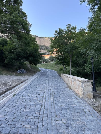 A paved walkway with cobblestone patterns curves through a lush, green park. The walkway is bordered by a stone wall, topped with a rough-hewn stone edge. A black lamppost stands sentinel near the wall. Beyond the walkway, dense trees with vibrant green foliage create a shaded canopy. In the distance, a dramatic, sun-drenched cliff face rises majestically under a clear blue sky. The scene appears to be set in the late afternoon or early evening, with the warm sunlight casting long shadows and highlighting the textures of the stone and vegetation. The overall atmosphere is one of tranquility and natural beauty, suggesting a peaceful outdoor space for a stroll or quiet contemplation in a mountainous or hilly region. There is no visible text in the image.