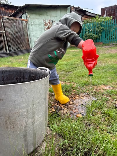 A young child, wearing a hooded sweatshirt with a crocodile graphic, grey pants, and yellow rain boots, is watering the ground with a red novelty watering can shaped like a cartoon animal. The child is standing in a grassy yard with some exposed dirt and pebbles. To the left of the child is a large, aluminum pot or bucket. In the background, there is a rustic wooden fence, a dilapidated wooden shed or outbuilding, and some overgrown vegetation. The sky is overcast. This scene appears to be in a rural or suburban setting, possibly a backyard, in Verkhnyaya Ury, Russia.