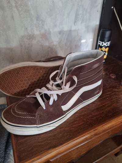This media file features a pair of brown Vans high-top sneakers with white laces and white side stripes. One shoe is prominently displayed, resting on a wooden surface. The sole of the shoe, with its distinctive waffle pattern, is visible. In the background, a portion of a black can with yellow and white text, possibly an aerosol product like AXE body spray, is visible. The overall scene appears to be indoors, with a light-colored, possibly marble or patterned surface beneath the shoes and a wooden table or shelf they are placed on. The lighting suggests it is daytime. There are no people present. The image does not provide specific cues for time of day or weather conditions, other than the general impression of daylight. The location context provided is Heusden-Zolder, Belgium, but this is not visually discernible from the image itself.