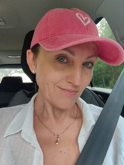 The image shows a woman wearing a pink baseball cap with a white heart design and a white collared shirt, sitting in a car. She is wearing a gold necklace with a cat pendant and a small stud earring in her visible ear. The woman has blue eyes and fair skin with freckles. She is looking towards the camera and smiling. The background shows the interior of a car, including a seatbelt and a headrest, and outside the window, trees are visible, suggesting a rural or suburban setting. The location is not specified as Хаценчыцкі сельскі Савет, Belarus.