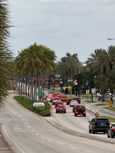A street scene in Bal Harbour Village, Florida, United States, shows a road with multiple lanes of traffic heading into the distance. The road is lined with numerous palm trees on both sides. Several vehicles are visible in traffic, including SUVs, sedans, and a van. Traffic signals and street signs are present, with a "SPEED LIMIT 30" sign clearly visible. A white oval sign with "Bal Harbour" written on it in cursive font is situated on a landscaped median. The sky is overcast. A person on a scooter is visible in the right lane of traffic.