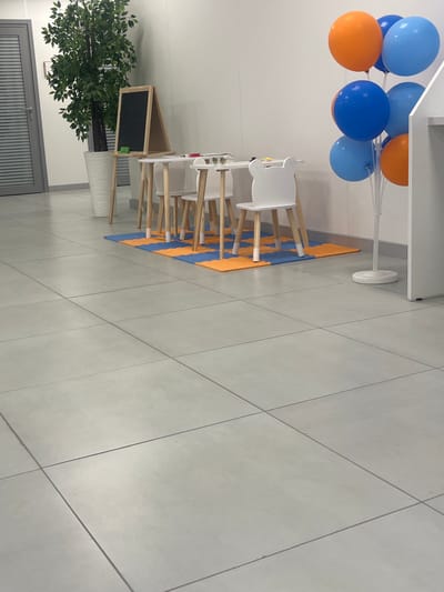 The image depicts a brightly lit interior space, possibly a classroom or play area, with a focus on a children's table and chairs. The scene is set against a clean, white wall and a grey tiled floor.
In the foreground and mid-ground, a light-colored table with wooden legs is visible, accompanied by several white chairs with a distinct, rounded, bear-ear-like design on the backrests. The chairs are arranged around the table, suggesting a setting for young children. Underneath the table and chairs, a mat with an alternating pattern of orange and blue squares is laid out on the floor.
To the left of the table, a large artificial potted plant with dark green leaves stands in a white decorative pot. Behind the plant, a grey door with horizontal slats is partially visible, possibly leading outside or to another room. Adjacent to the plant and the table, a wooden easel with a blackboard is positioned. A green marker is resting on the ledge of the blackboard.
To the right of the table, a cluster of colorful balloons is tethered to a white stand. The balloons are a mix of bright orange and various shades of blue, creating a festive atmosphere. A portion of a white shelving unit or partition is also visible on the far right of the image.
The lighting appears to be a mix of natural and artificial, creating a well-illuminated environment. The overall impression is one of a tidy and welcoming space, likely designed for children's activities.
No people are present in the image. The setting does not provide specific visual cues to pinpoint its exact location within Novotroitsk, Russia.